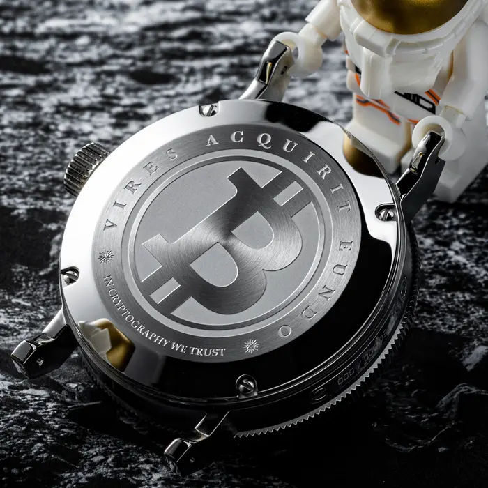 A CRYPTOMATIC Jetsetter caseback engraved with the Bitcoin symbol and the motto Vires Acquirit Eundo, resting on a textured black surface.