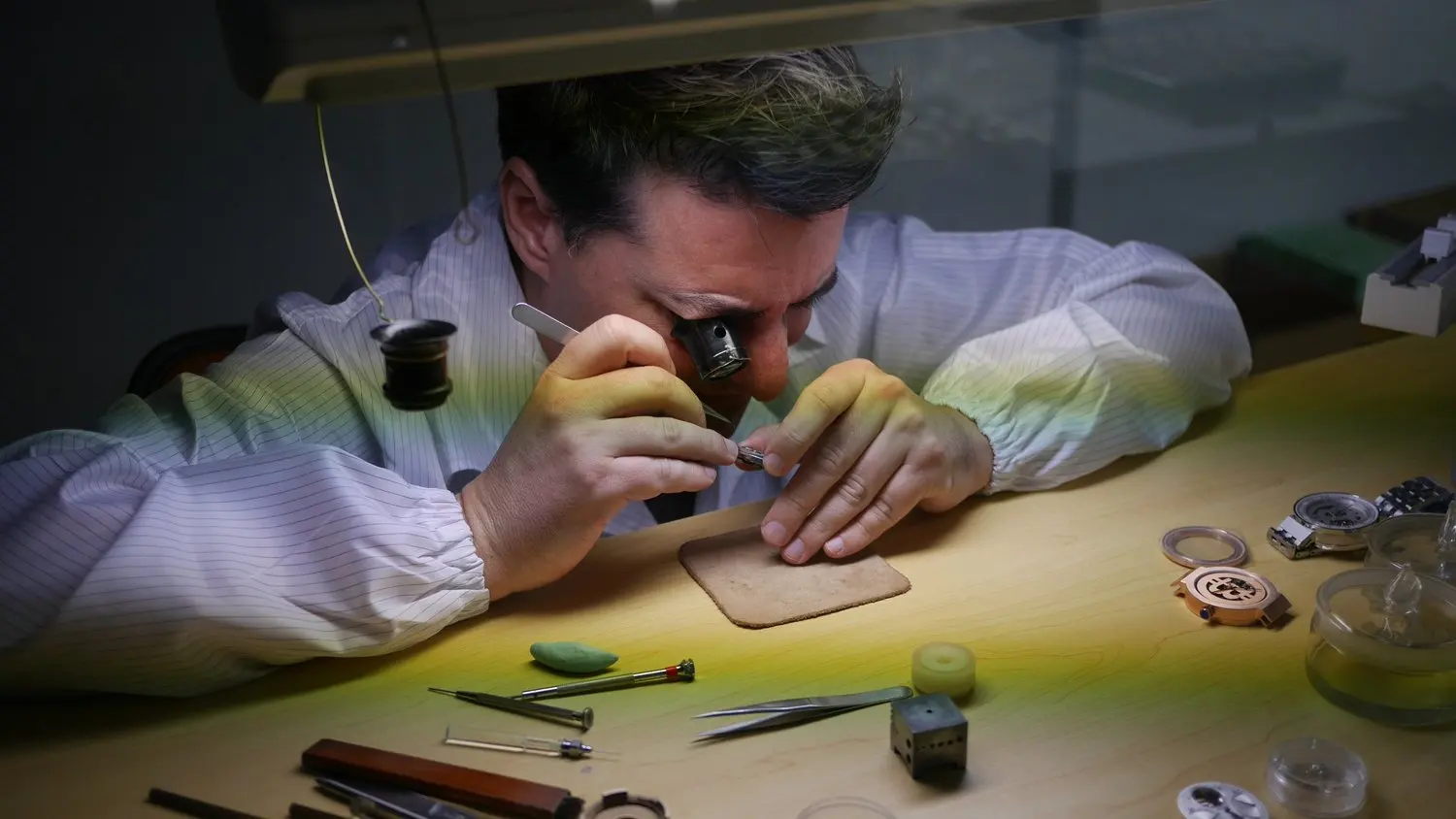 A watchmaker working meticulously on a small watch part under a magnifying lens at a workbench with various watchmaking tools.