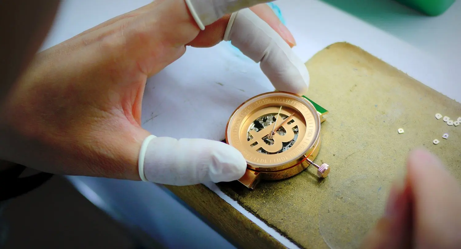 Close-up of a person's hands wearing white gloves assembling a watch with a gold-colored face and exposed internal mechanism on a workbench.