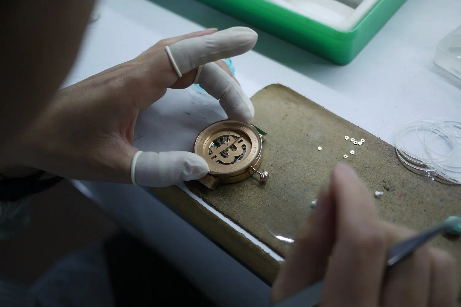 Person wearing white gloves working on a watch in a workshop, with small watch parts and tools on the work surface.