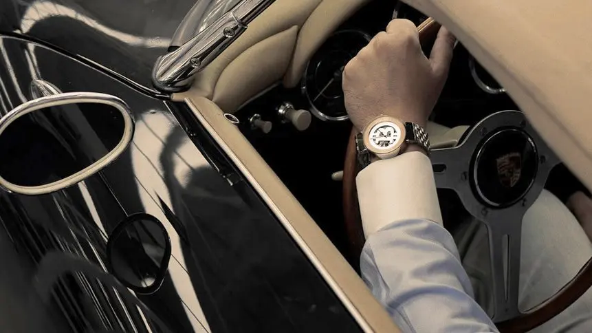 Person driving a classic black car, wearing a beige watch and a light gray jacket, with a wooden steering wheel and vintage interior.