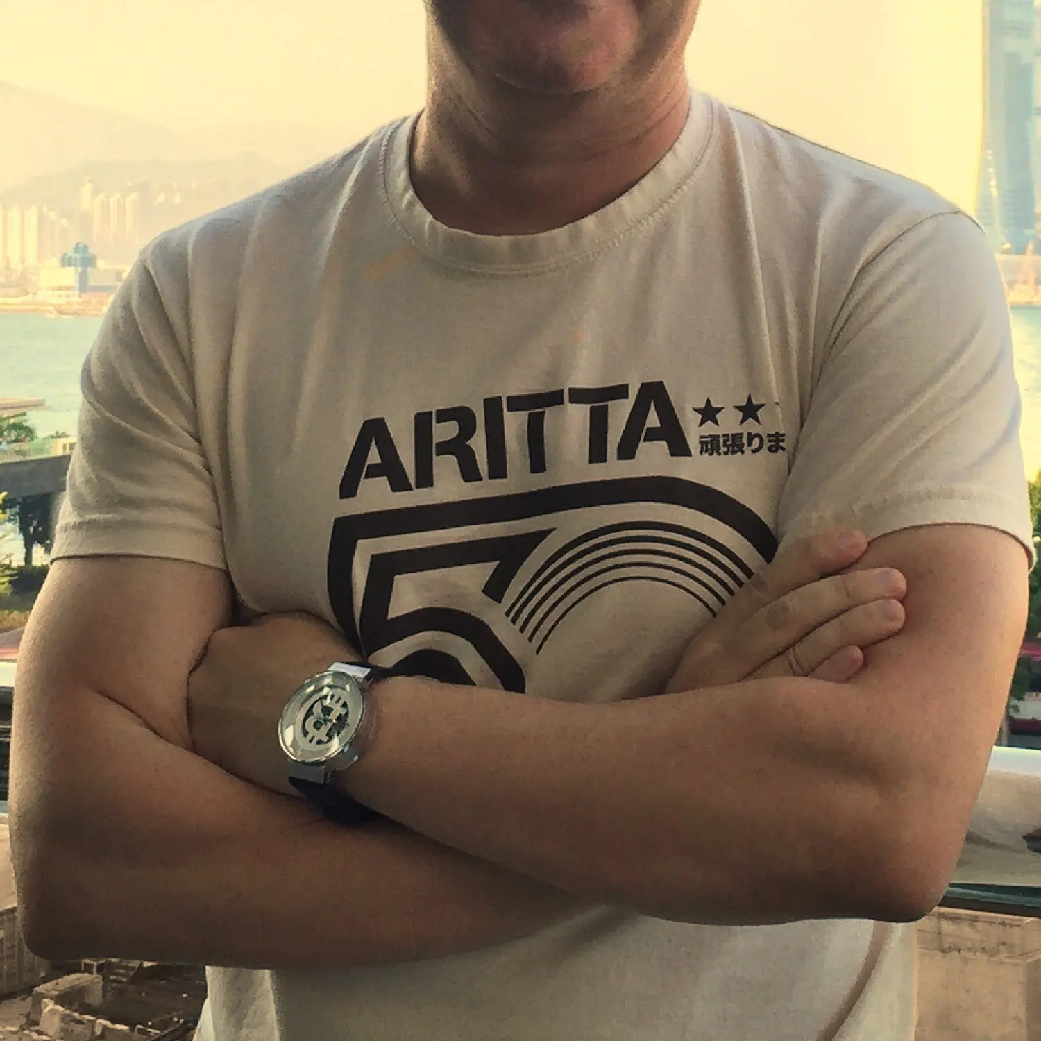 Close-up of a person with crossed arms wearing a Peter Pan collar T-shirt with a graphic design and partial Japanese text, standing in front of a window overlooking a cityscape with tall buildings and a river.