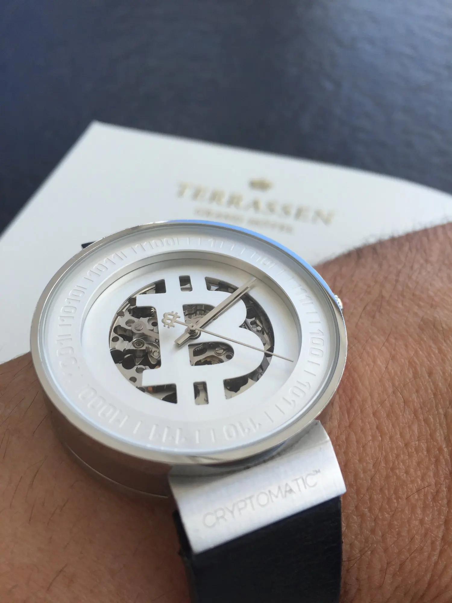 A wristwatch with a white face and a silver bezel, featuring the Bitcoin symbol and mechanical gears visible through the open center.