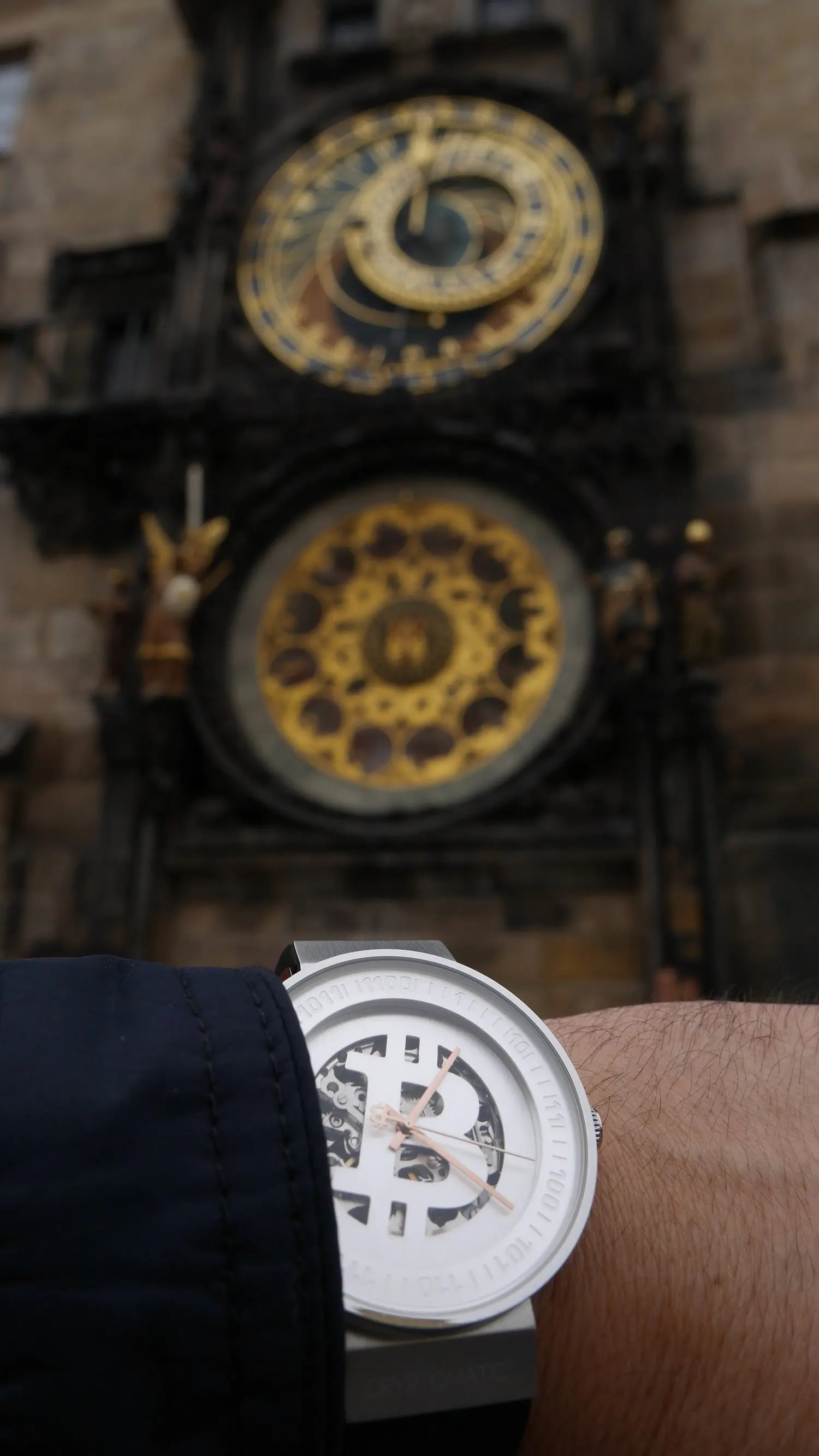 A wristwatch with a Bitcoin symbol on the face, worn on a person's arm, with a blurred historical clock tower in the background.