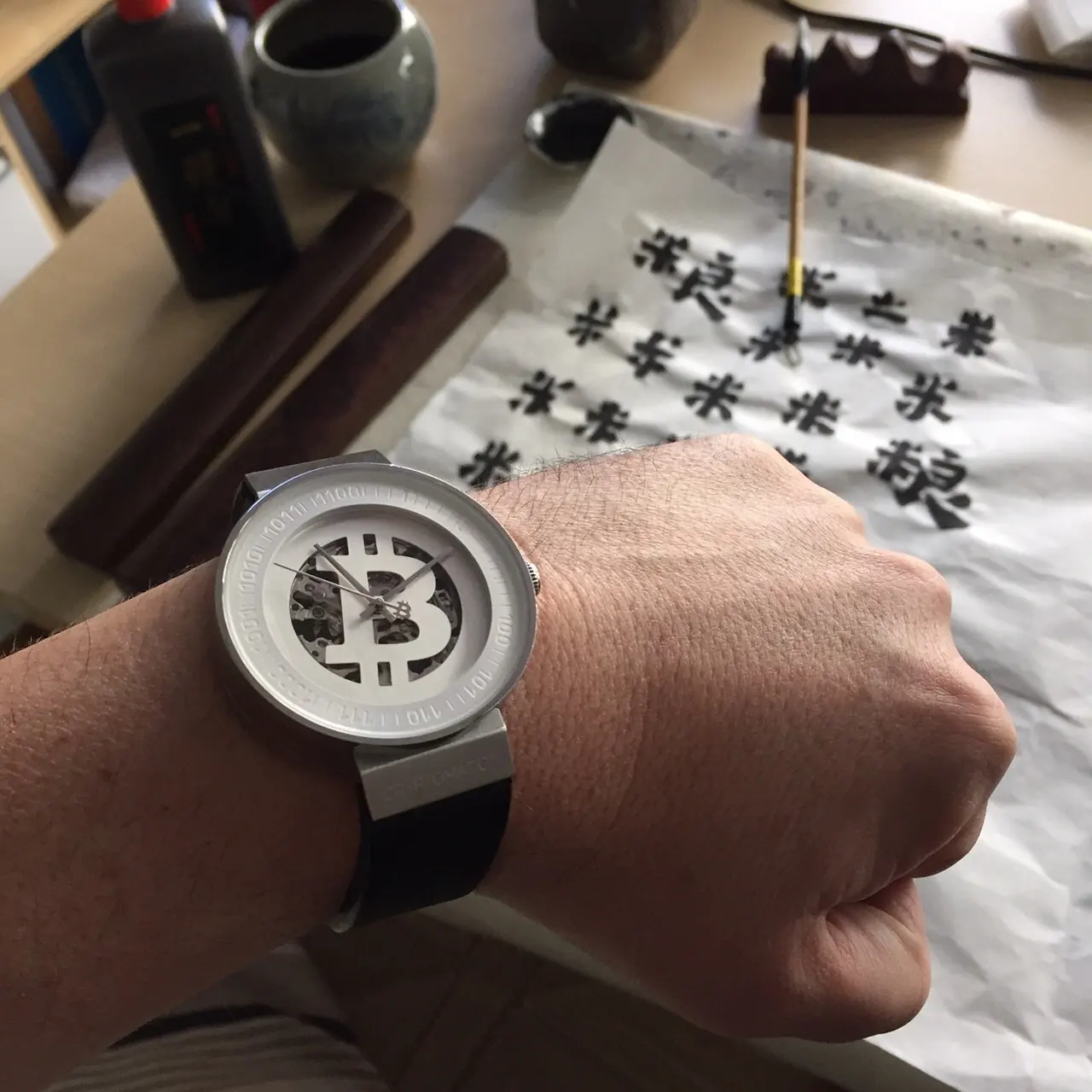 A person's left wrist wearing a watch with a large Bitcoin symbol on the face, observing a workspace with paper featuring multiple Bitcoin symbols, ink brush, and artist tools.