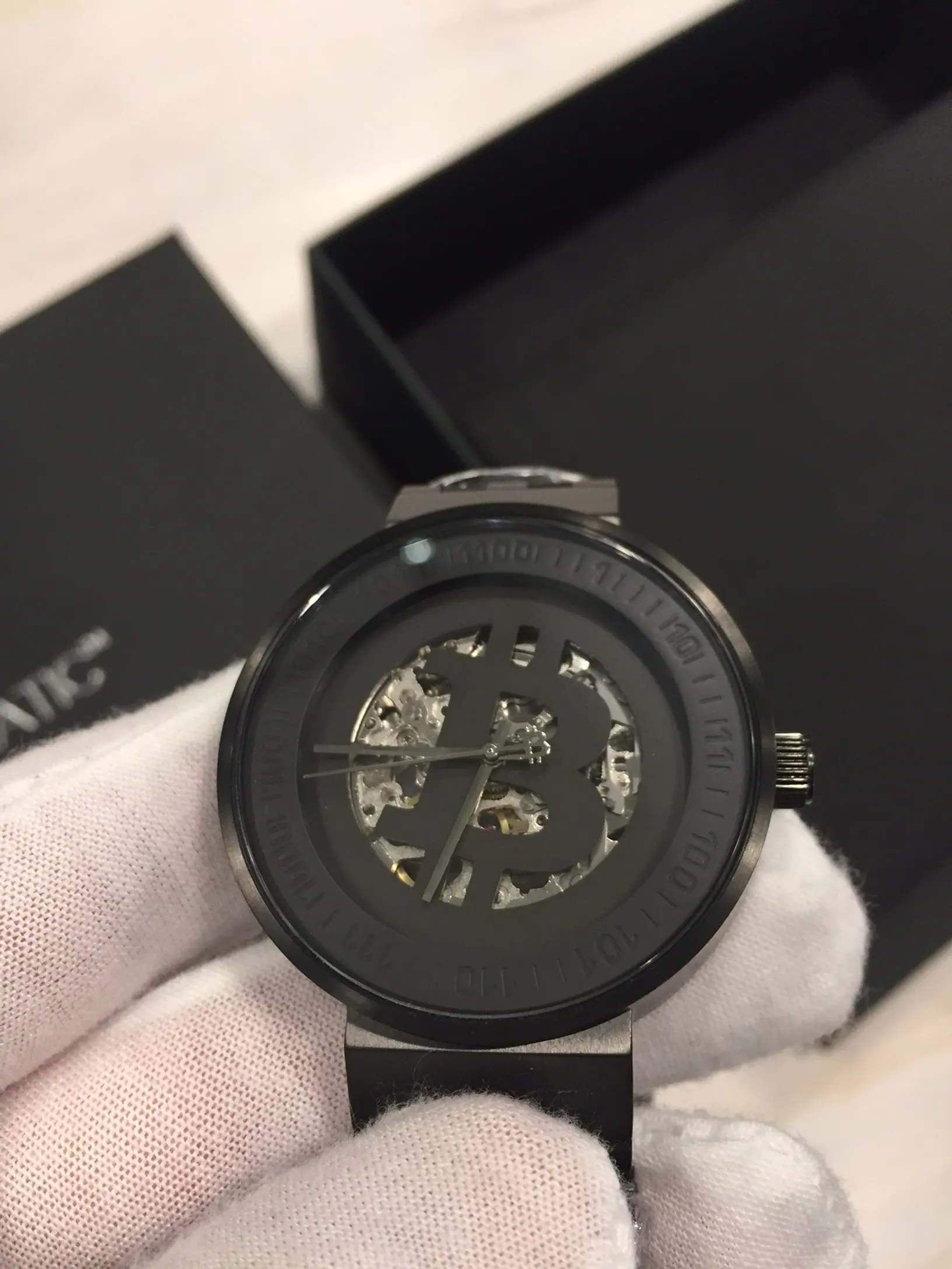 Black wristwatch with a skeleton dial and Bitcoin symbol in the center, held by a gloved hand with a black box in the background.