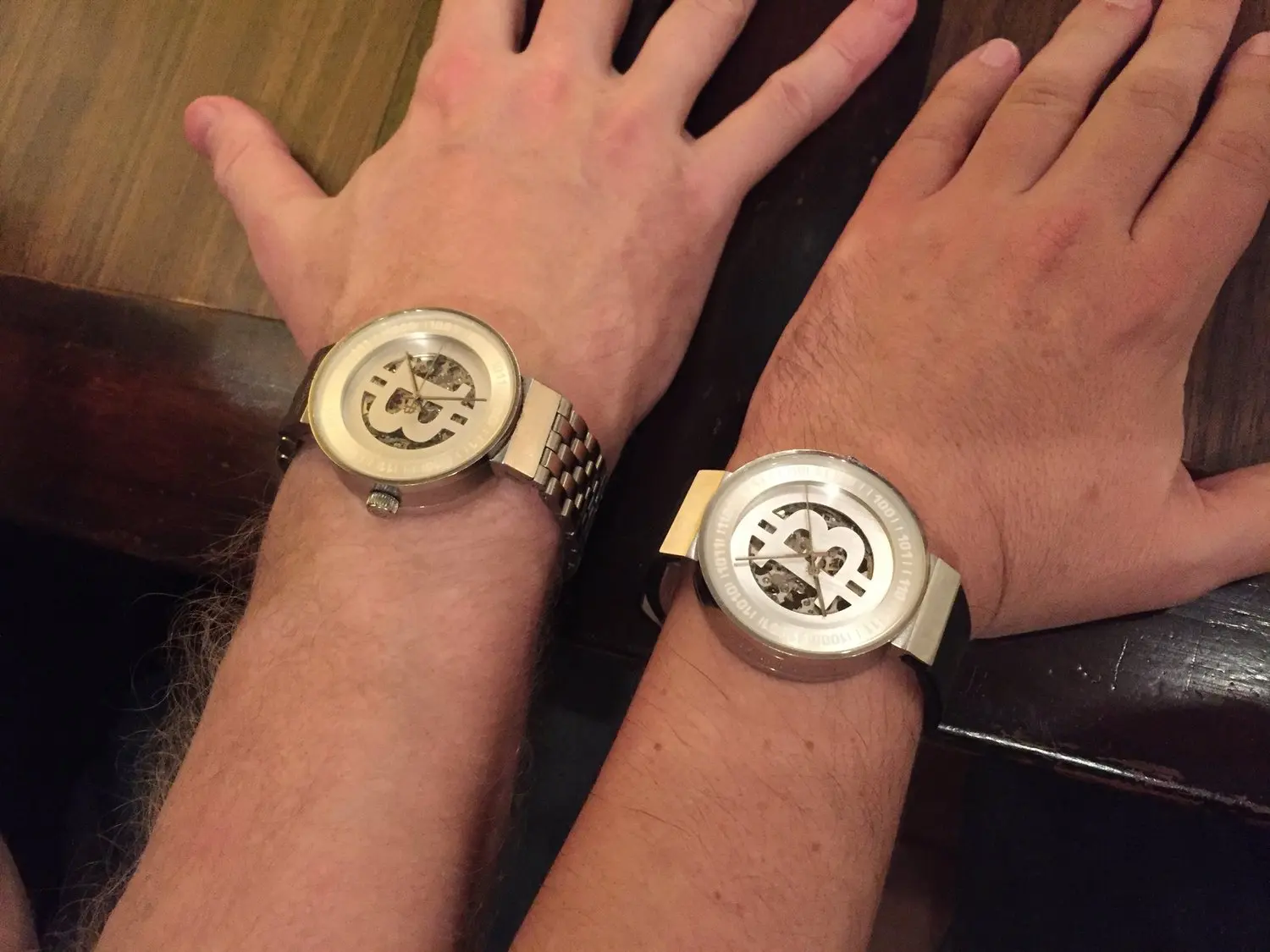 Two people wearing wristwatches with open skeleton dials showing the inner mechanisms, resting their hands on a wooden surface.