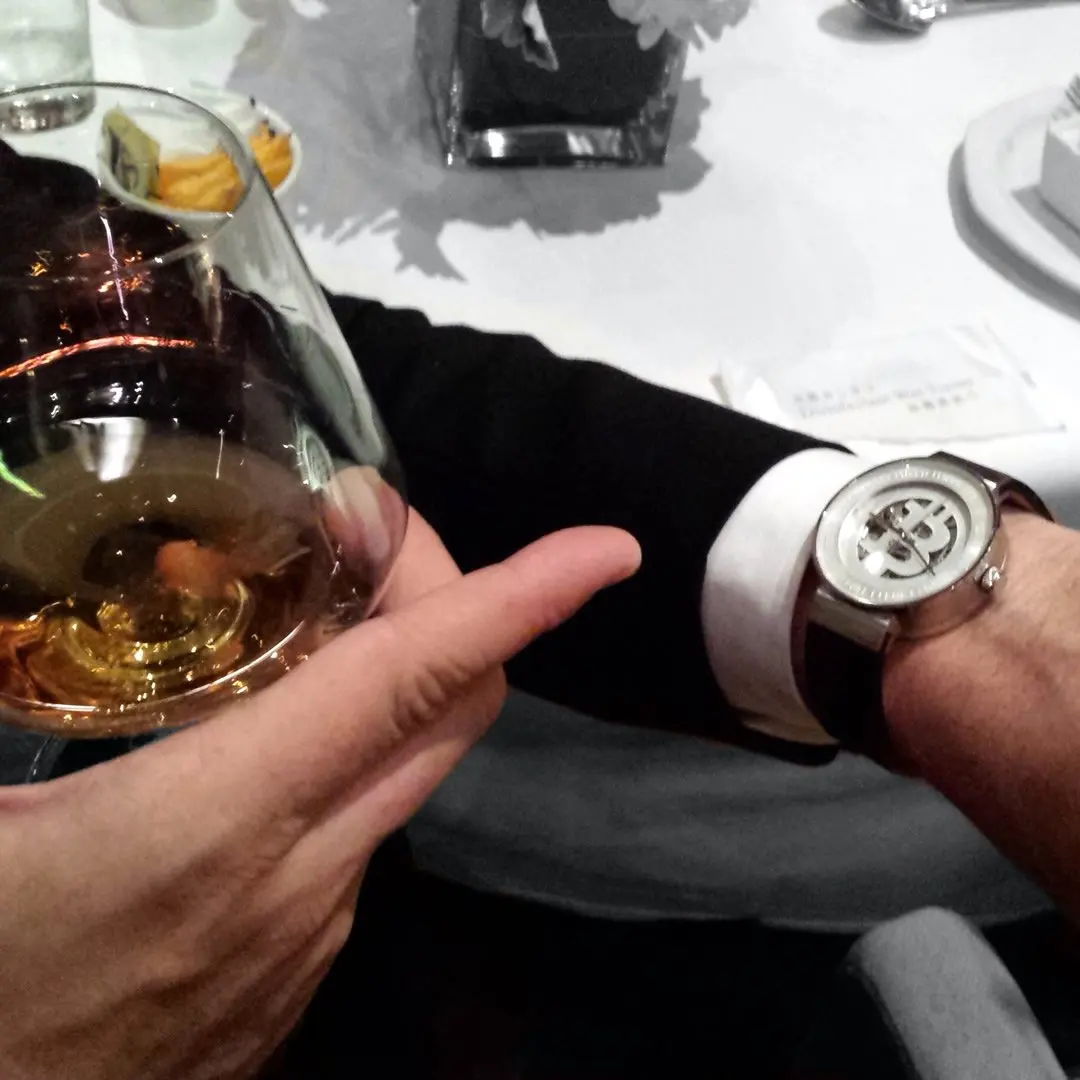 Person holding a glass of white wine at a table with a black-strap bitcoin wristwatch visible, in a dining setting.