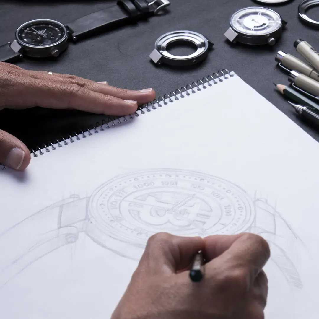 A person sketches a bitcoin watch design on a spiral notebook, surrounded by watch faces, pens and watch parts on a dark surface.