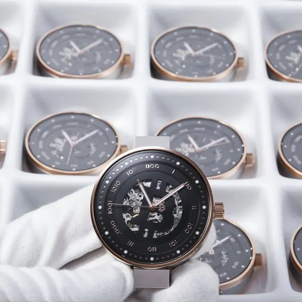 A person wearing a white glove holding a black and gold bitcoin wristwatch, with several similar watches in white boxes in the background.