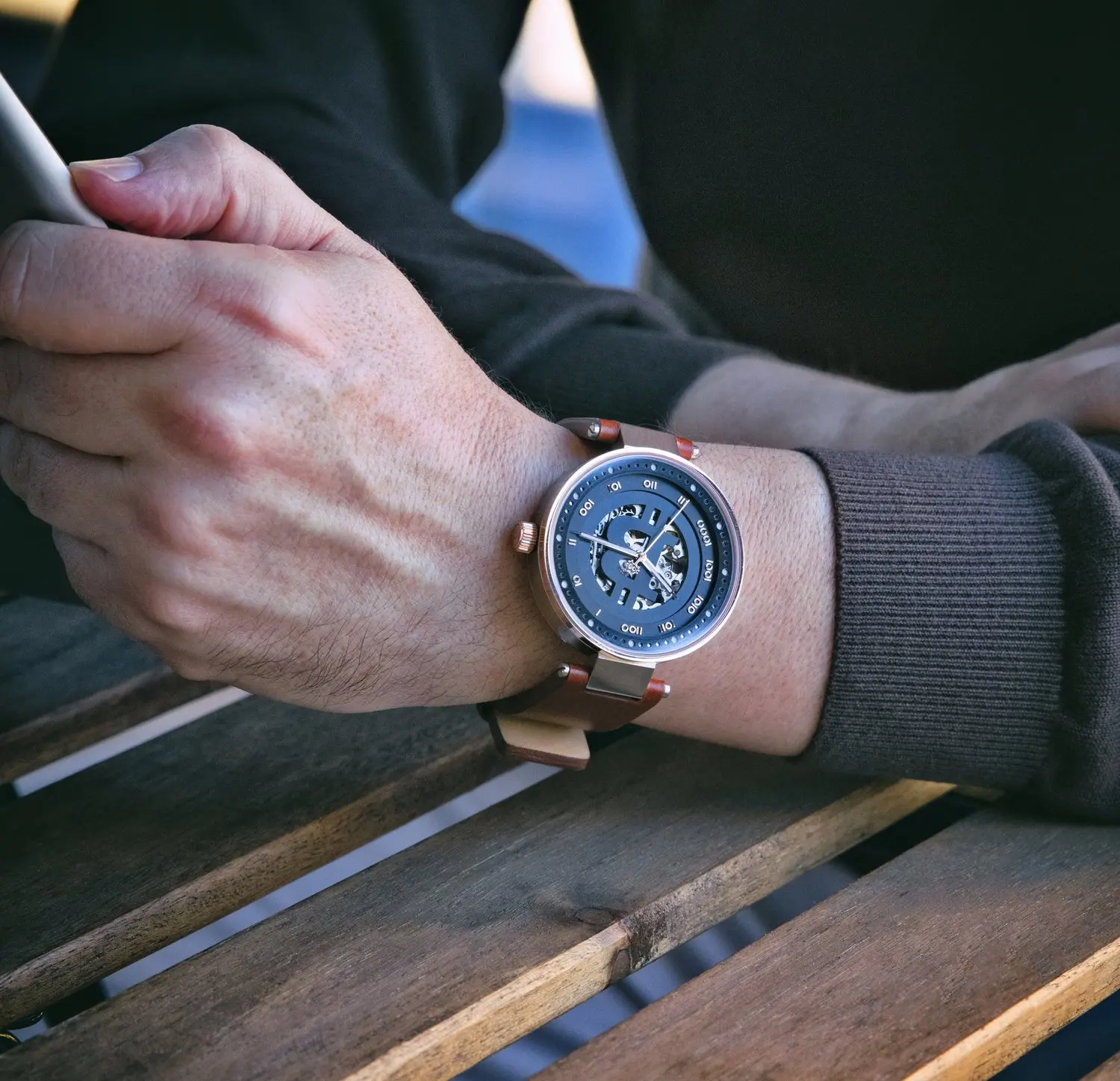 A person's left wrist wearing a stylish bitcoin wristwatch with a dark face and a brown leather strap, resting on a wooden surface while holding a smartphone.