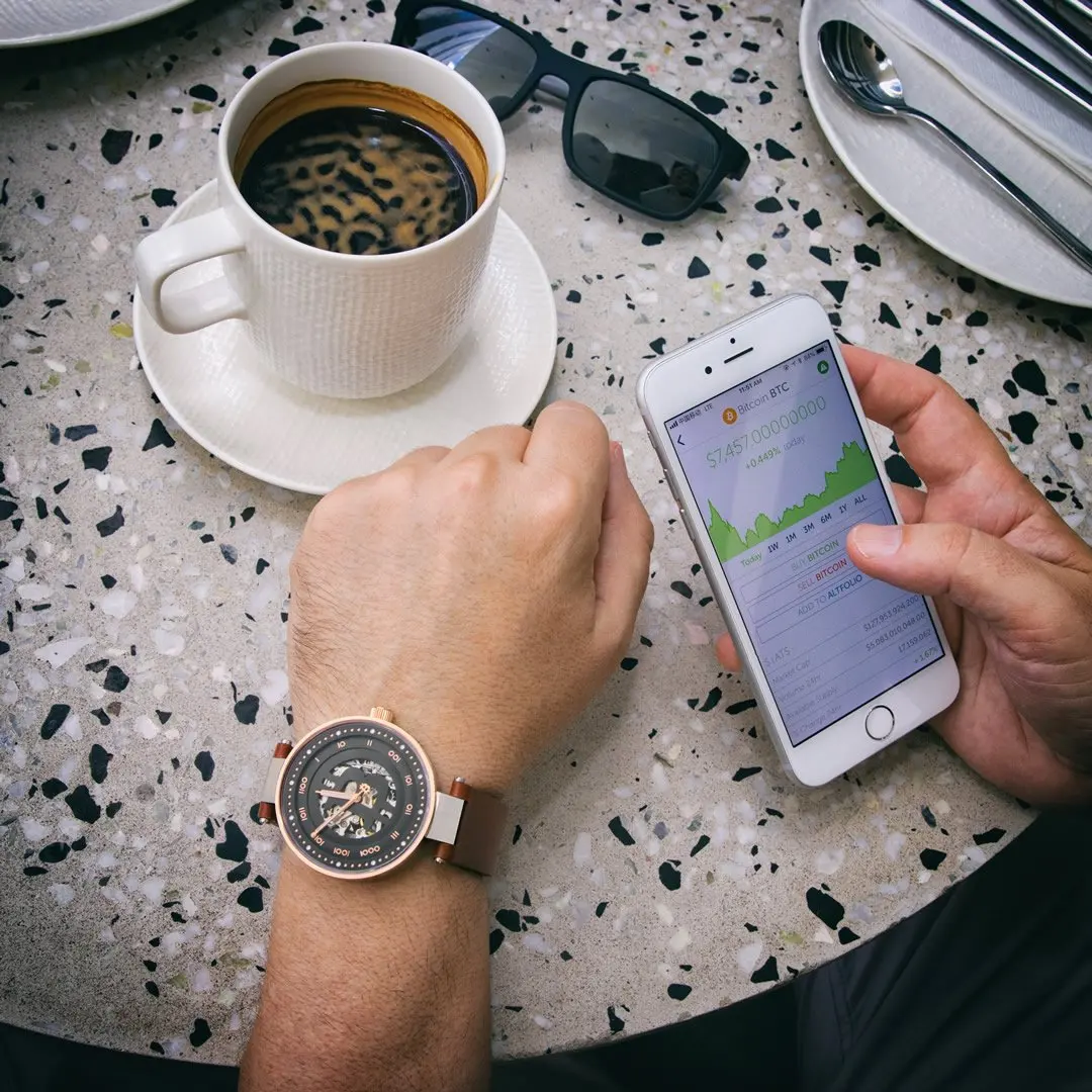 Person using a smartphone displaying a Bitcoin price chart, on a speckled tabletop with a cup of coffee, sunglasses and a wristwatch.