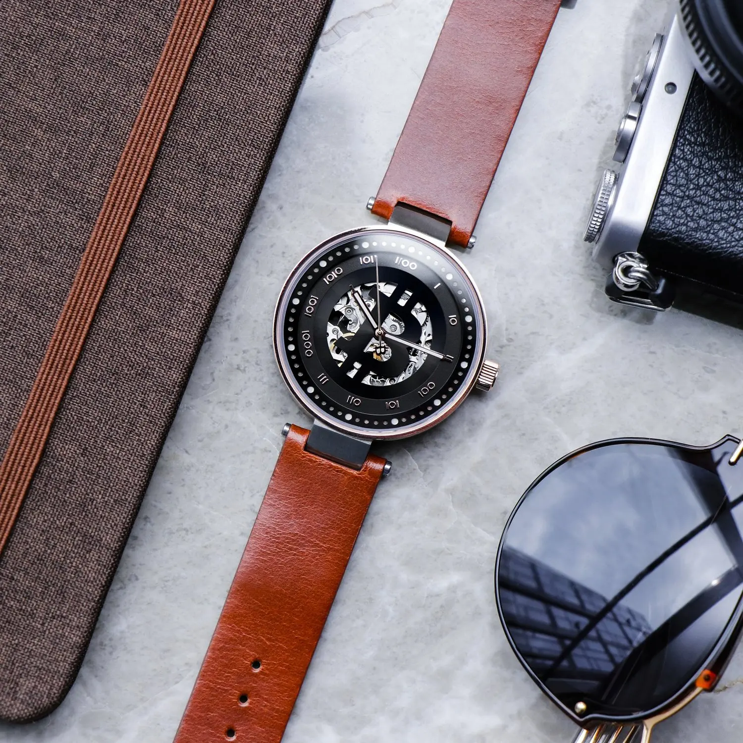 A bitcoin wristwatch with a brown leather strap and a black and silver dial, placed on a light gray marble surface alongside a brown textured case, a black camera and sunglasses.