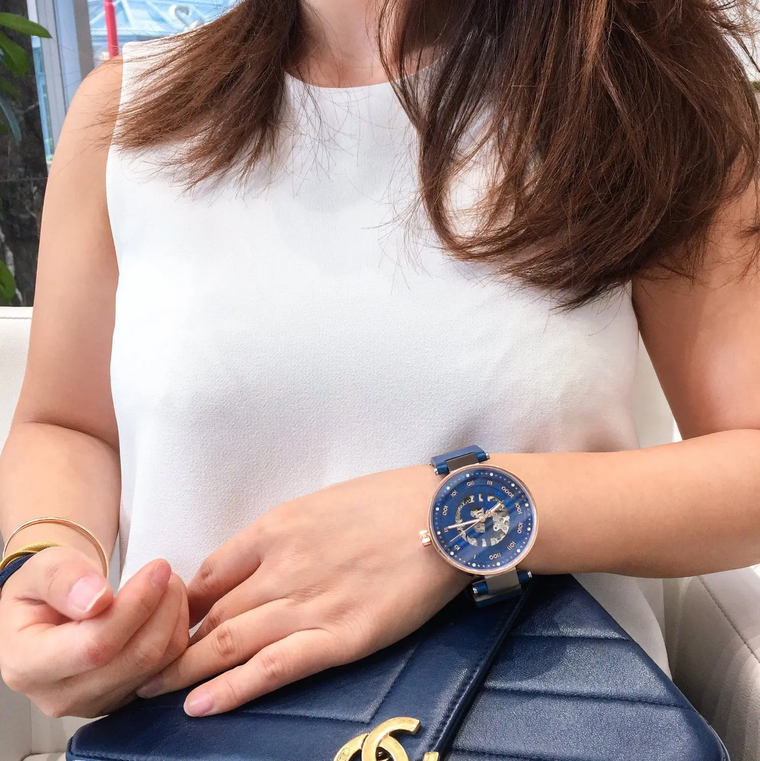 A woman wearing a white sleeveless top, a blue bitcoin watch and a gold bracelet, sitting indoors.