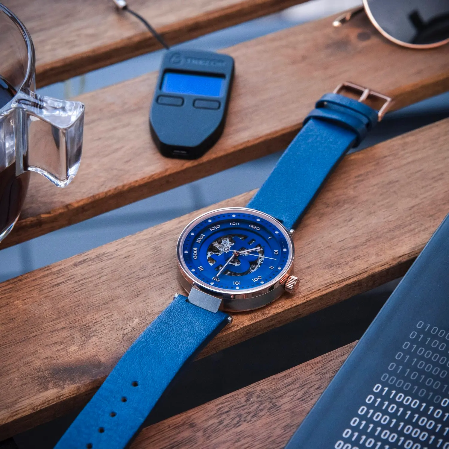 A blue bitcoin wristwatch with a partially transparent face showing internal gears, placed on a wooden surface with sunglasses and a sheet of binary code.