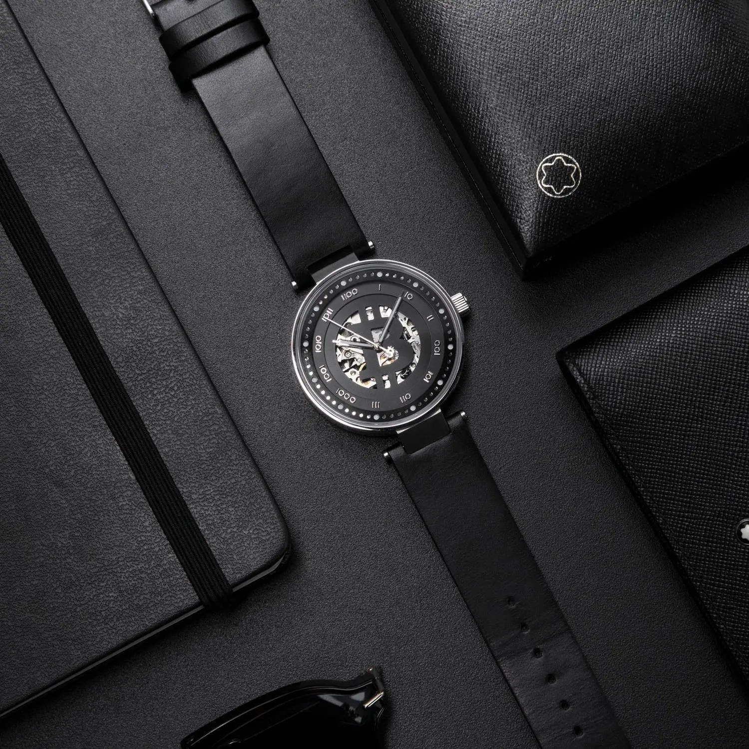 A black bitcoin wristwatch with a visible mechanical movement, placed on a black textured surface alongside notebooks and a leather wallet.