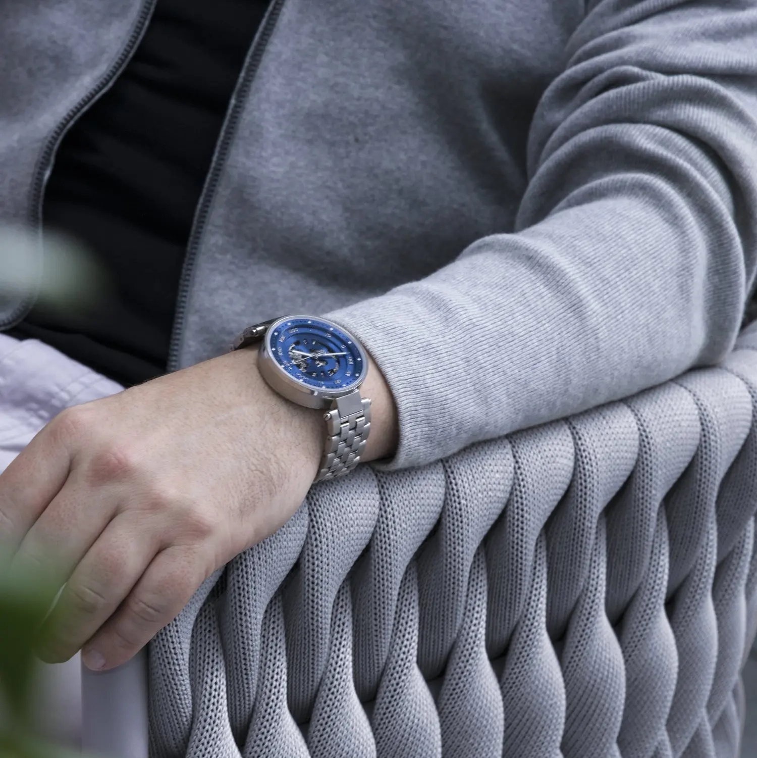 A person's left arm resting on a woven chair, wearing a silver bitcoin wristwatch with a blue face and a gray hoodie.
