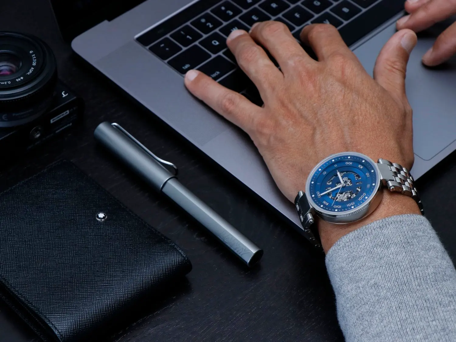 A person's wrist wearing a silver and blue bitcoin wristwatch while typing on a laptop keyboard.