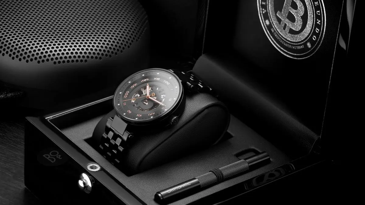 A luxury black wristwatch with a metal band displayed on a watch cushion inside a black box, alongside a pen.