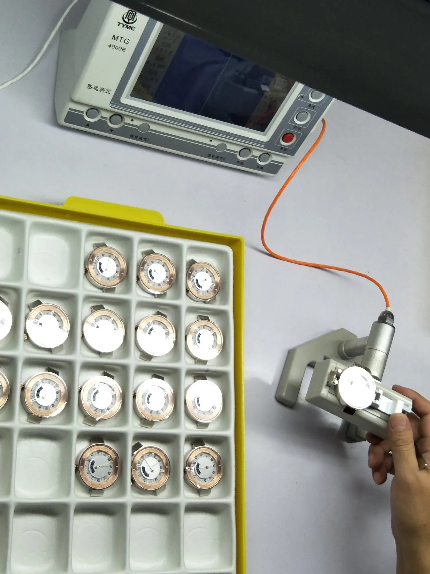 A person holding a device connected to an orange wire, testing small round dials arranged in a yellow tray.