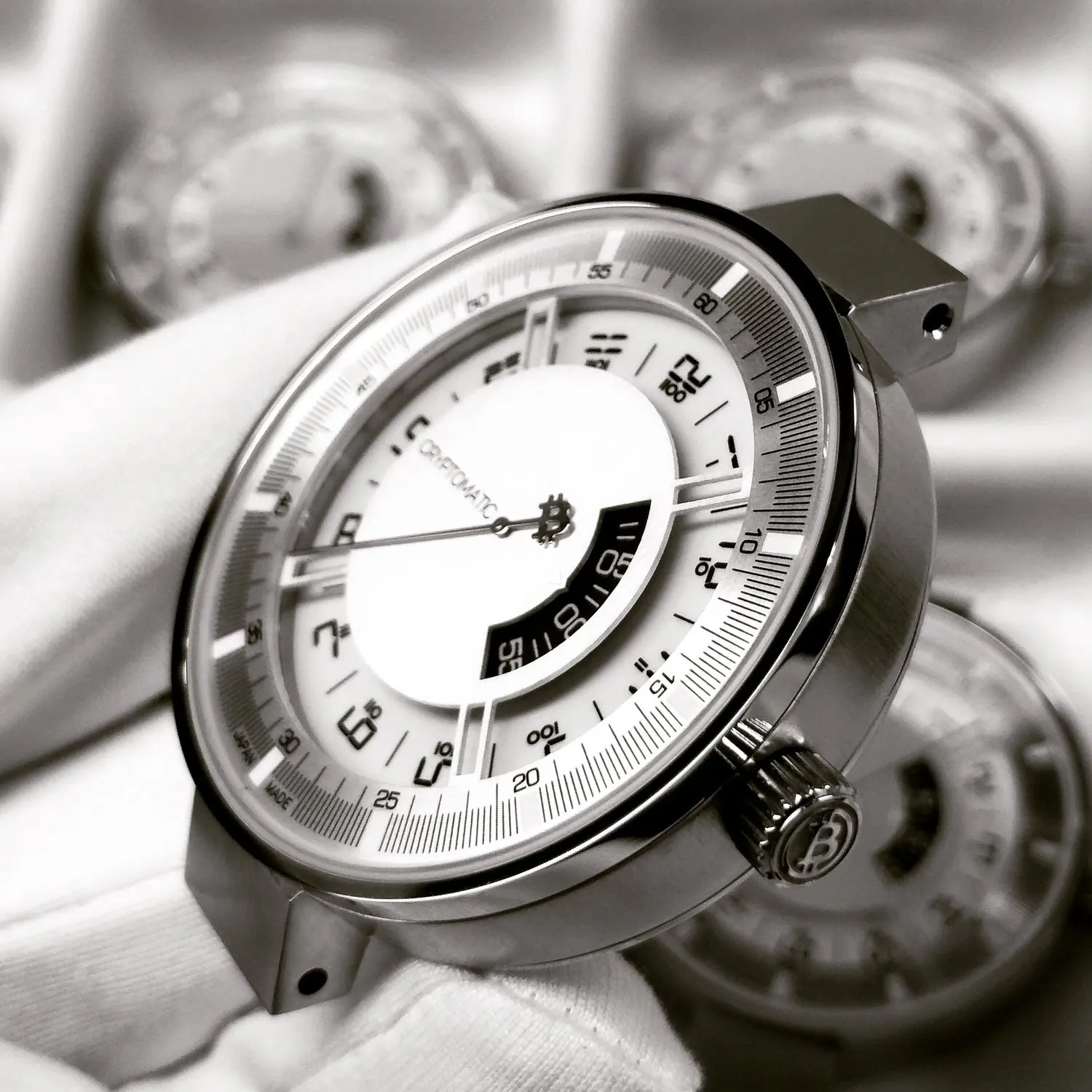 Close-up of a luxury bitcoin wristwatch with a white face, black and white details, and a metal case, with additional watches blurred in the background.