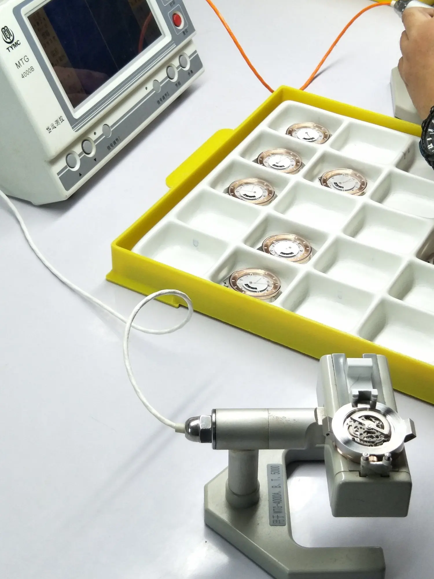 Watchmaking workspace with watch dial components organised in a yellow tray, a watchmaker's tool and an electronic testing device.
