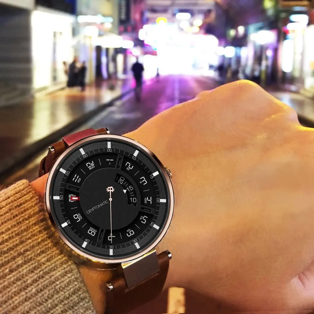 A person's left wrist with a modern bitcoin watch displaying the time and Bitcoin symbol, held up against a blurred city street at night.