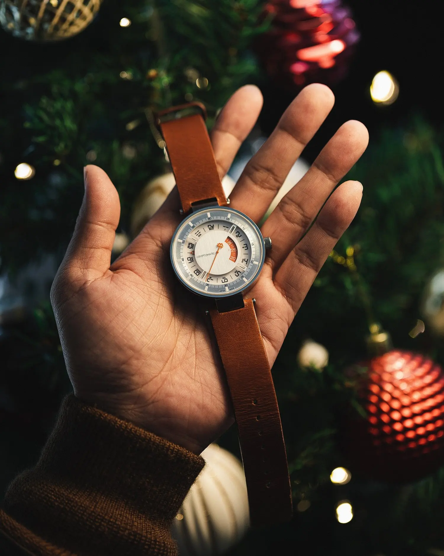A person's hand holding a bitcoin wristwatch with a brown leather strap in front of a decorated Christmas tree.